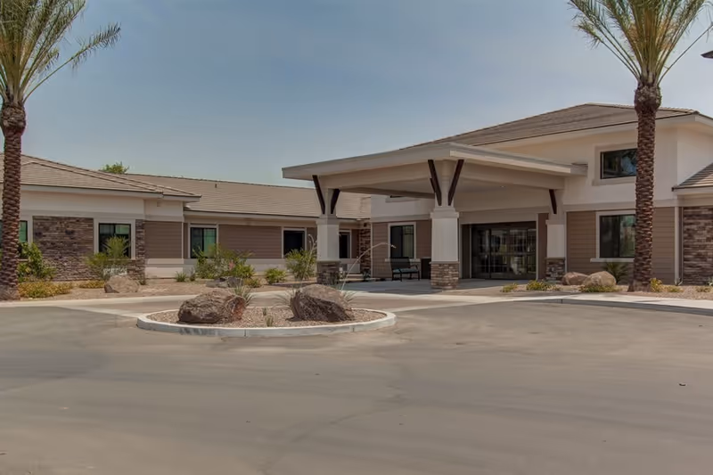Single-story senior living building entrance with a covered porte-cochere, circular driveway, and palm trees.
