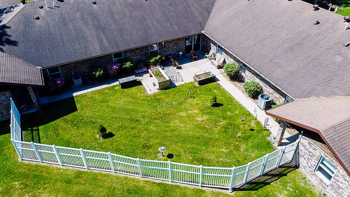 Aerial view of a fenced-in grassy courtyard area surrounded by a single-story brick building with a brown roof. The courtyard features a concrete patio with chairs, raised garden beds, small shrubs, and decorative garden stakes.