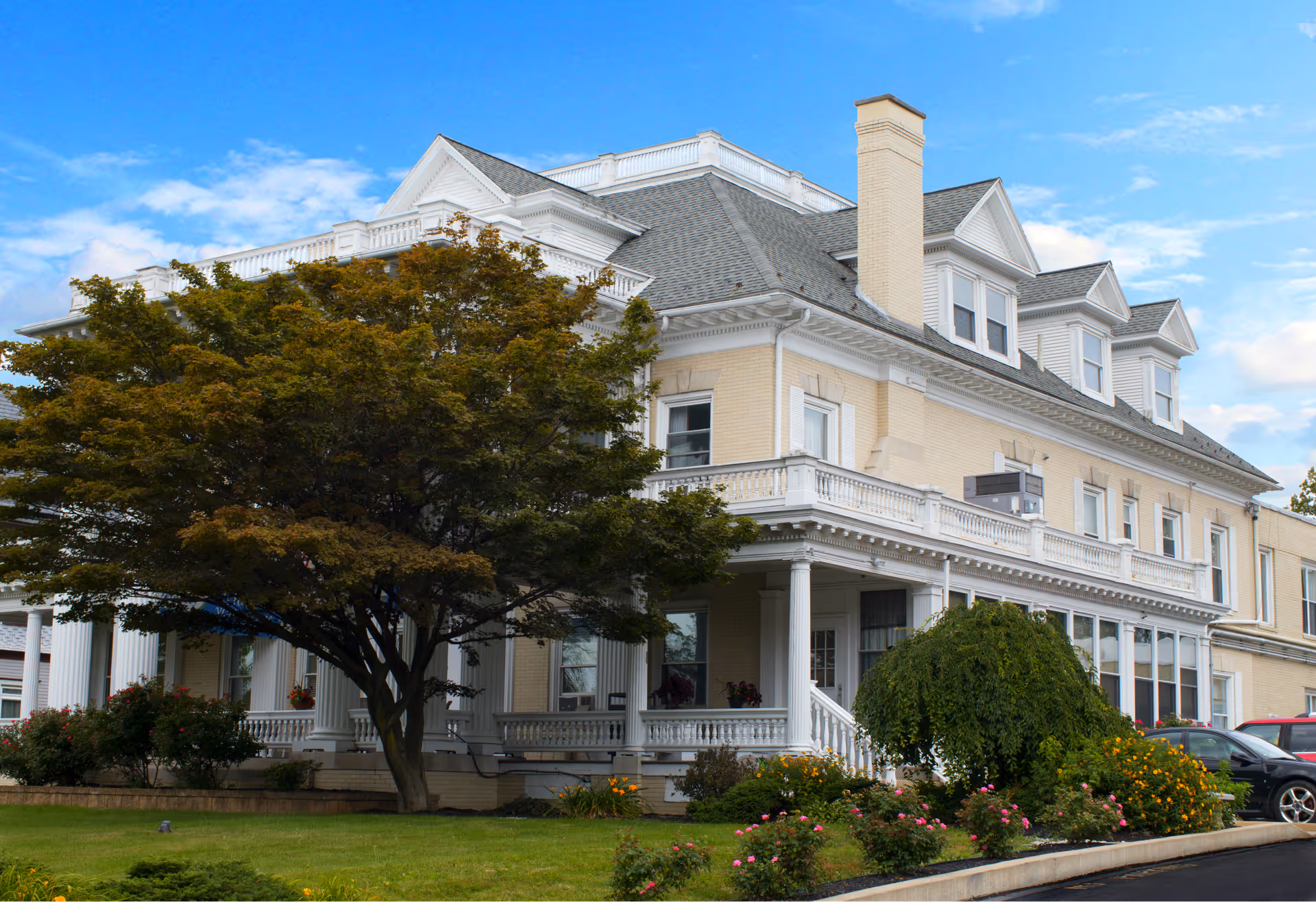 Exterior view of a large, elegant, multi-story building with a light yellow brick facade, white trim, and a gray shingled roof. The building has a porch with white columns and railings, surrounded by well-maintained landscaping including a large tree, bushes, and flowers. Several cars are parked near the building under a partly cloudy blue sky.