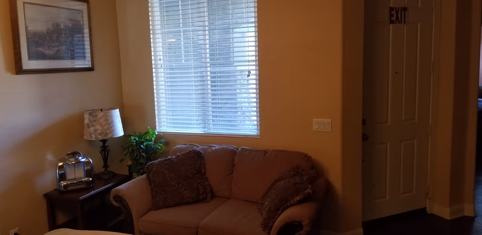 A cozy living room area with a brown cushioned loveseat adorned with two decorative pillows. Next to the loveseat is a small wooden side table holding a lamp with a patterned shade and a vintage-style radio. A green potted plant is placed beside the table. A window with white blinds allows natural light to enter the room. On the wall above the side table hangs a framed picture. To the right, there is a white door with an EXIT sign above it.
