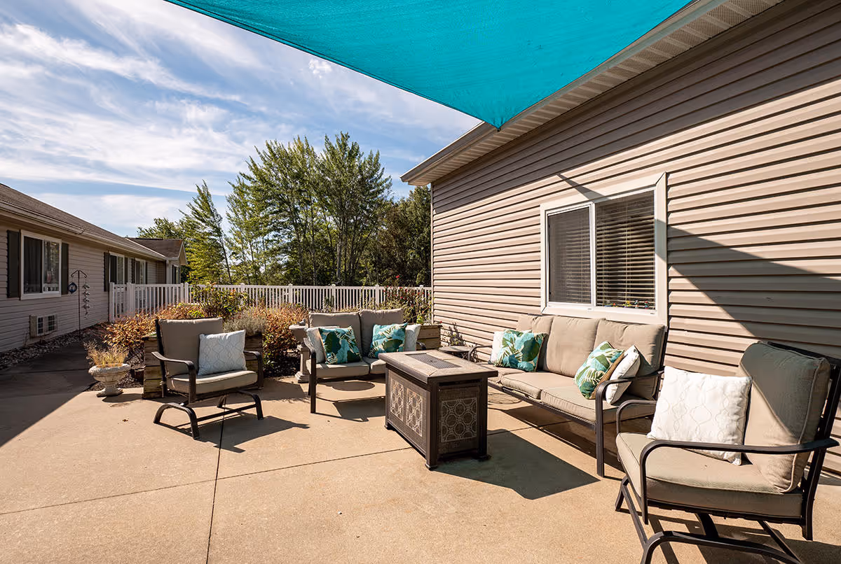 Outdoor patio area at Cedar Creek of Washington with cushioned seating including a sofa and chairs arranged around a central fire pit table. The patio is shaded by a large teal umbrella, and there are plants and trees in the background under a partly cloudy sky.