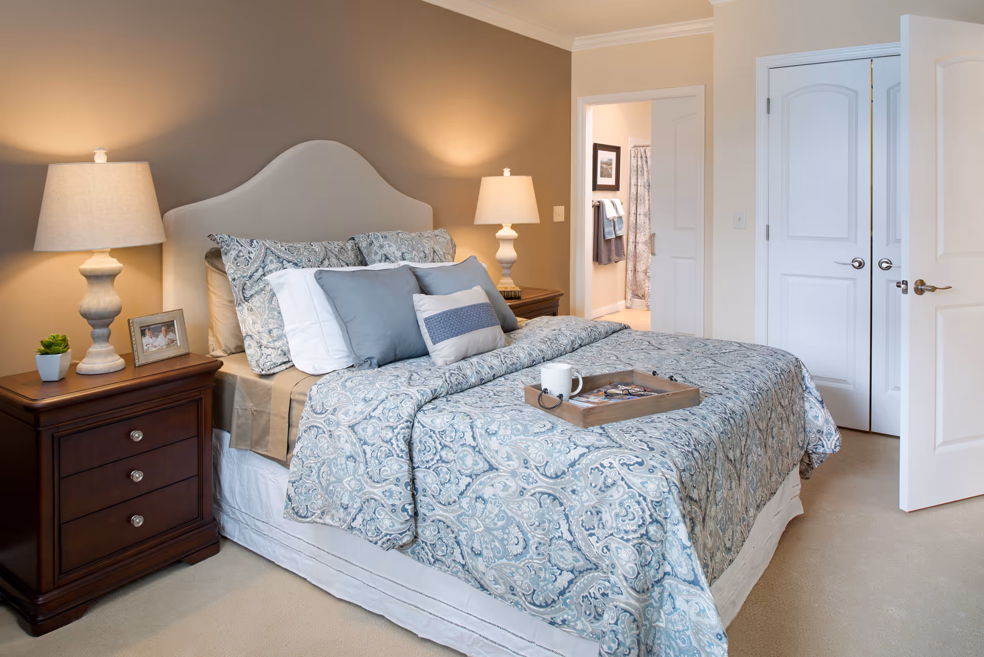 A neatly made bed with patterned blue and white bedding and multiple pillows in a bedroom. There are two bedside tables with lamps, a small plant, and a framed photo. An open door reveals a bathroom with towels hanging and a shower curtain visible.