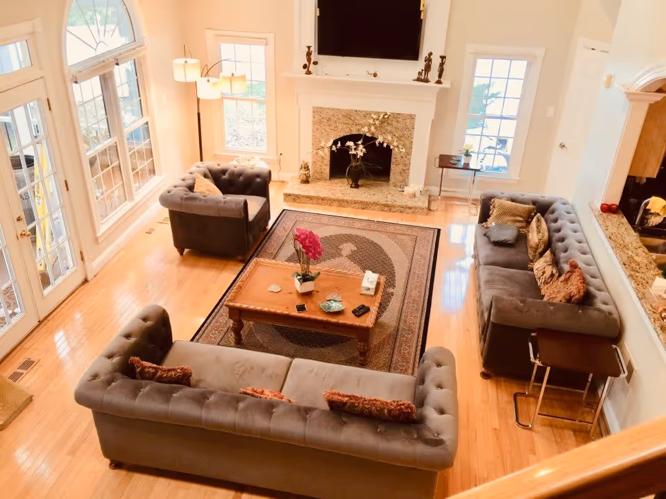 Bright living room with three gray tufted sofas arranged around a wooden coffee table on a patterned rug. The room features hardwood floors, a stone fireplace with decorative items on the mantel, large windows, and a glass door letting in natural light. A floor lamp stands in the corner, and the kitchen counter with granite top is partially visible on the right.
