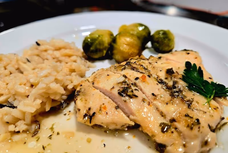 A close-up of a plate with cooked rice, roasted Brussels sprouts, and herb-seasoned sliced chicken breast garnished with a sprig of parsley.