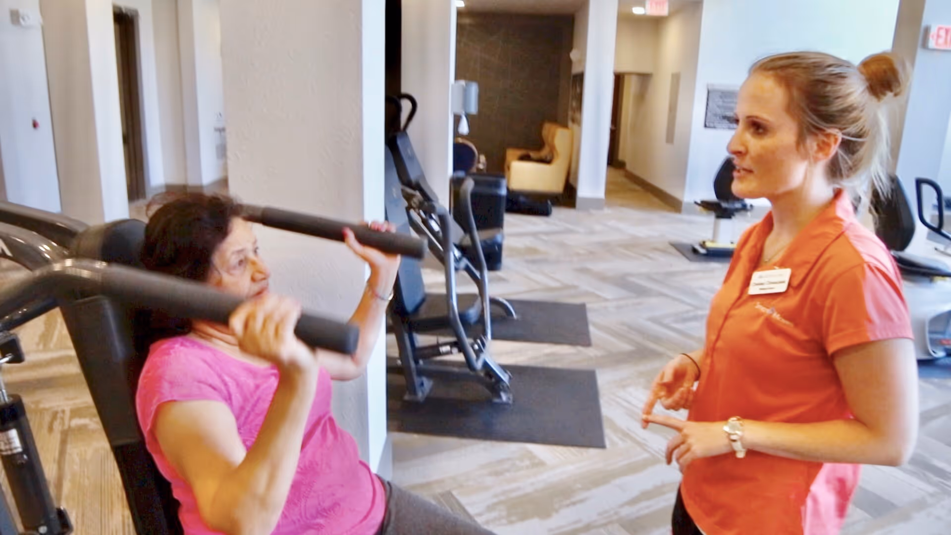 An elderly woman in a pink shirt is using a chest press machine in a fitness area while a female staff member in an orange shirt with a name tag is talking to her and providing guidance.