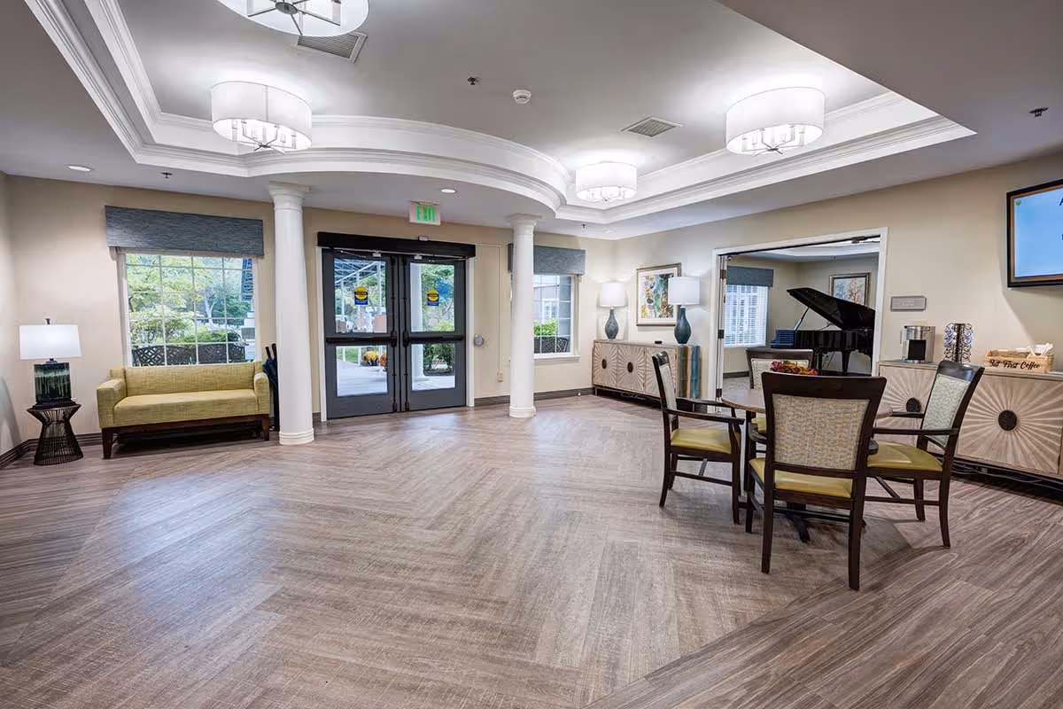 A spacious, well-lit common area in a senior living facility featuring a seating arrangement with four chairs around a small table, a green loveseat near a window, decorative lamps, and a grand piano in an adjacent room. The room has wood-patterned flooring, white walls, and ceiling lights with a tray ceiling design. Double glass doors lead outside, and there are large windows allowing natural light to enter.