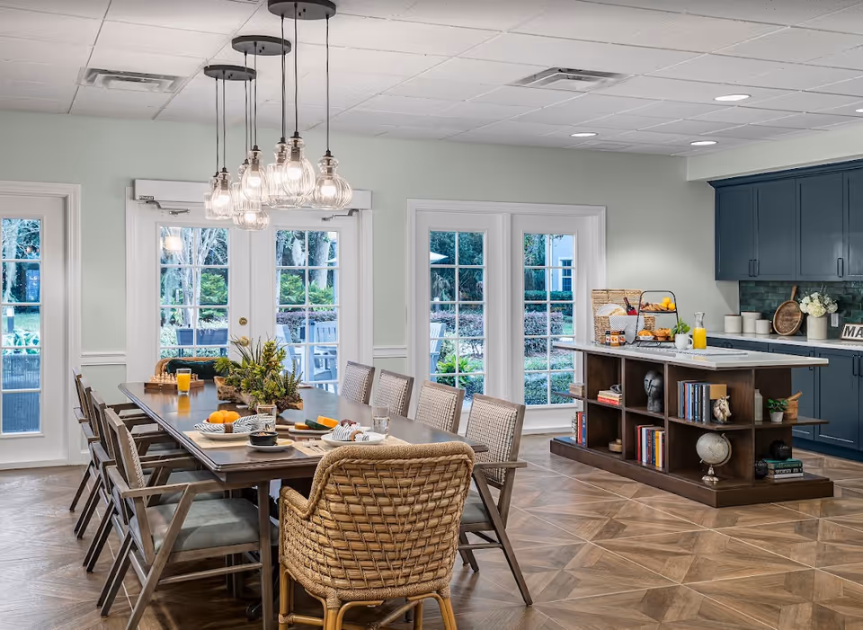 Bright communal dining area with a long wooden table surrounded by wicker chairs, hanging pendant lights, and a kitchen island beside French doors to an outdoor patio.