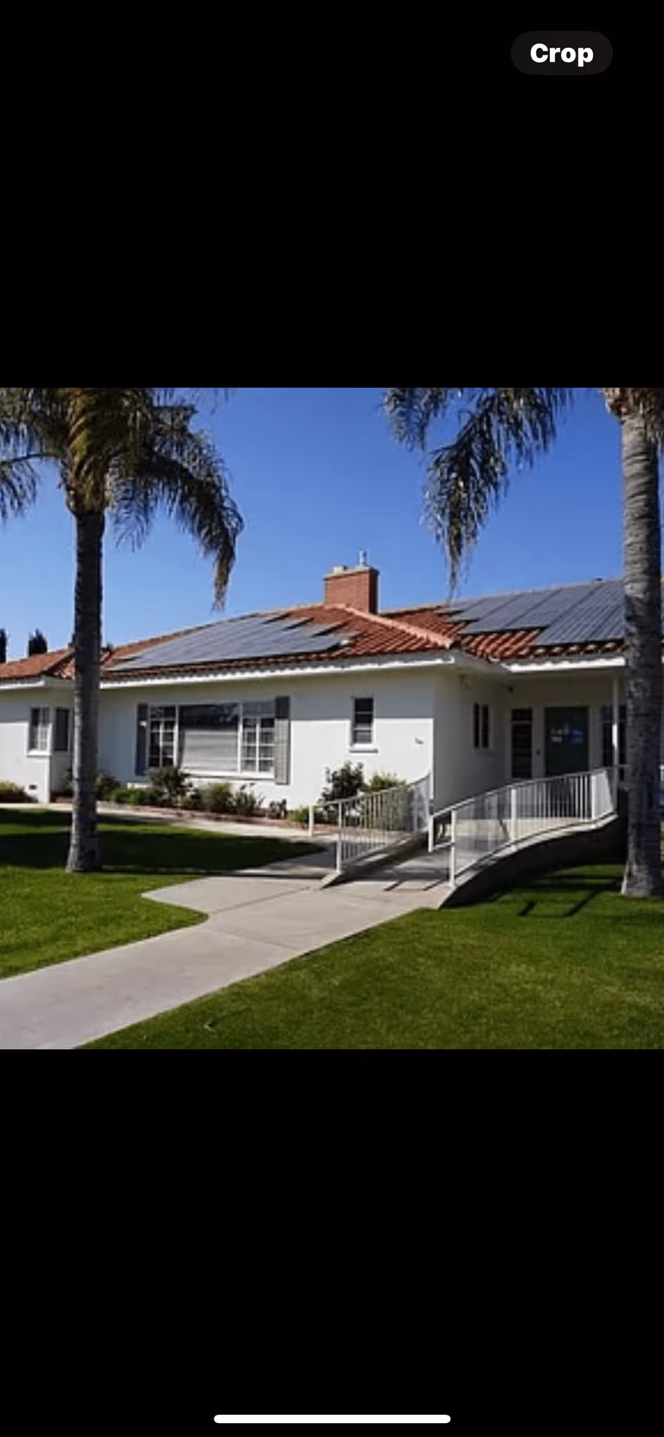 Exterior view of a single-story building with a red-tiled roof and solar panels, surrounded by green grass and palm trees, with a concrete walkway and wheelchair ramp leading to the entrance.