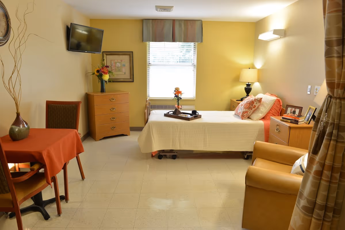 A cozy bedroom in a senior living facility with a single bed covered in a white quilt and orange pillows. There is a wooden dresser with a vase of flowers and a framed picture on the wall above it. A small table with two chairs and a red tablecloth is on the left side. A flat-screen TV is mounted on the wall above the dresser. A comfortable armchair is positioned near the bed, and a window with blinds and a valance lets in natural light. The room has warm yellow walls and tiled flooring.