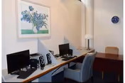 A small office or workspace with two computer monitors, speakers, and a keyboard on a desk. There are two chairs in front of the desk, a framed floral artwork on the wall, a table lamp, and a clock on the wall.