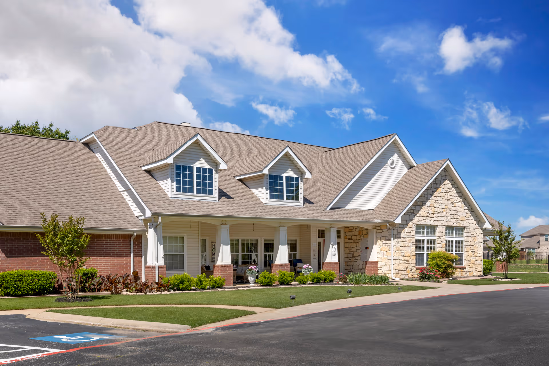 Exterior view of a single-story senior living facility building with a combination of brick, stone, and siding. The building features a covered porch with columns, several windows, and a well-maintained lawn with shrubs and flowers. The sky is blue with some clouds.