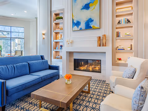 Bright seating area with a blue sofa, armchairs, a wooden coffee table, and a fireplace with built-in shelves and artwork above.