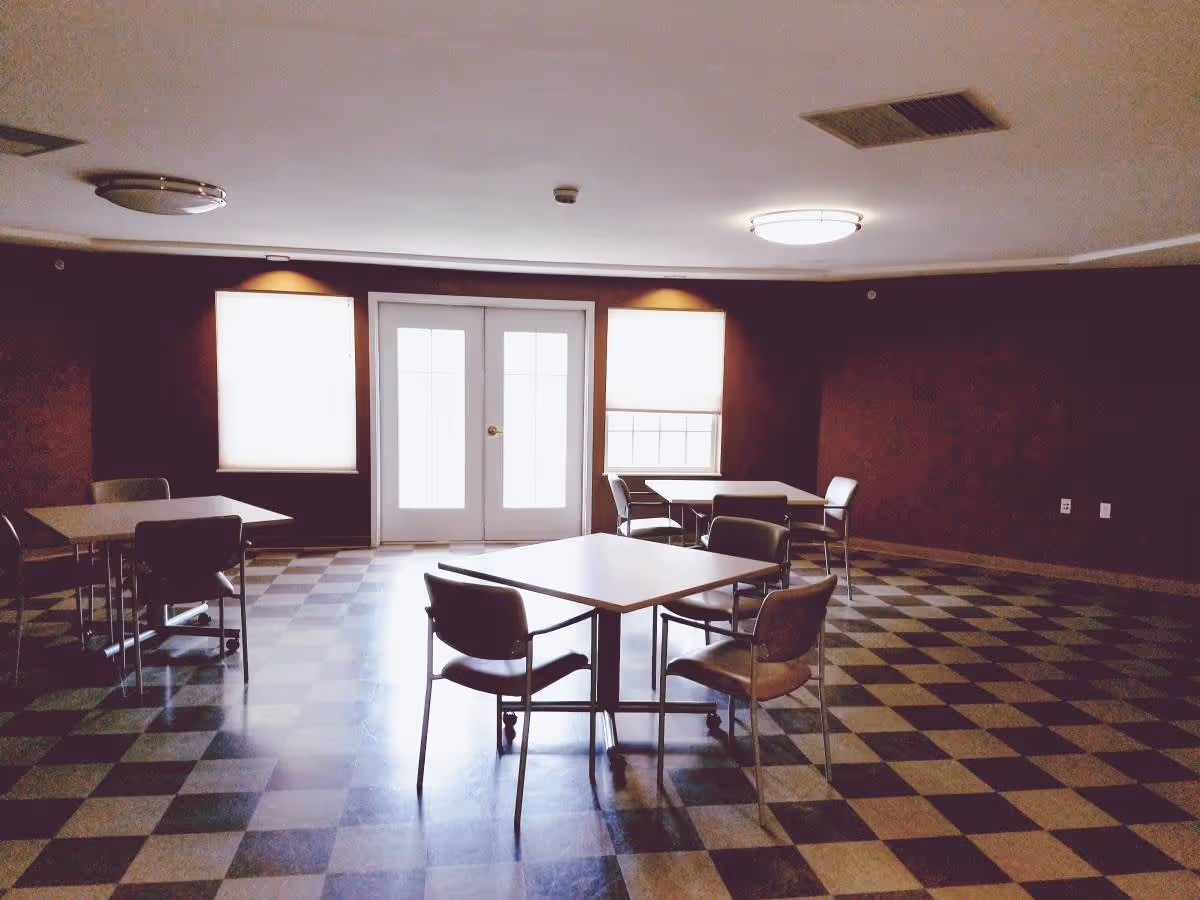 Interior room with checkered floor tiles, three square tables each surrounded by four chairs, two windows with blinds, and a set of double glass doors in the background.