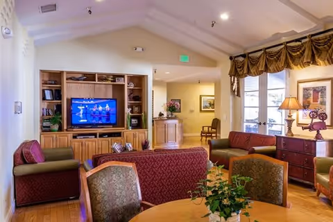 A cozy living room area in a senior living facility with a wooden entertainment center holding a television, several armchairs and sofas with patterned upholstery, a round table with chairs, and a sideboard with a lamp and framed artwork. The room has wooden flooring, a high ceiling with recessed lighting, and large windows with decorative curtains.