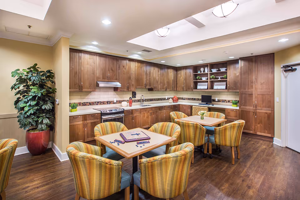 A bright and spacious kitchen and dining area with wooden cabinets, a stove, and a sink. There are two square tables with six striped upholstered chairs around them. One table has a Scrabble game set up. The room has wooden flooring, recessed lighting, and a large potted plant in a red pot in the corner.