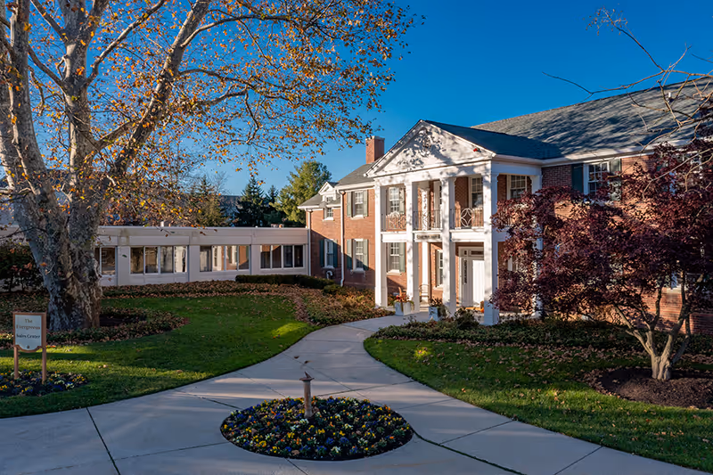 Exterior view of a senior living facility named The Evergreens, featuring a large brick building with white columns at the entrance, surrounded by green lawns, trees with autumn leaves, and a circular flower bed in the foreground under a clear blue sky.