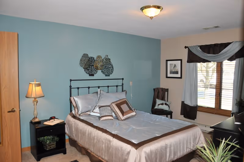 A neatly made bed with light blue and brown bedding in a bedroom with a light blue accent wall. There is a black metal headboard, a nightstand with a lamp and a book, a chair with a pillow, a window with curtains, and a framed picture on the wall.