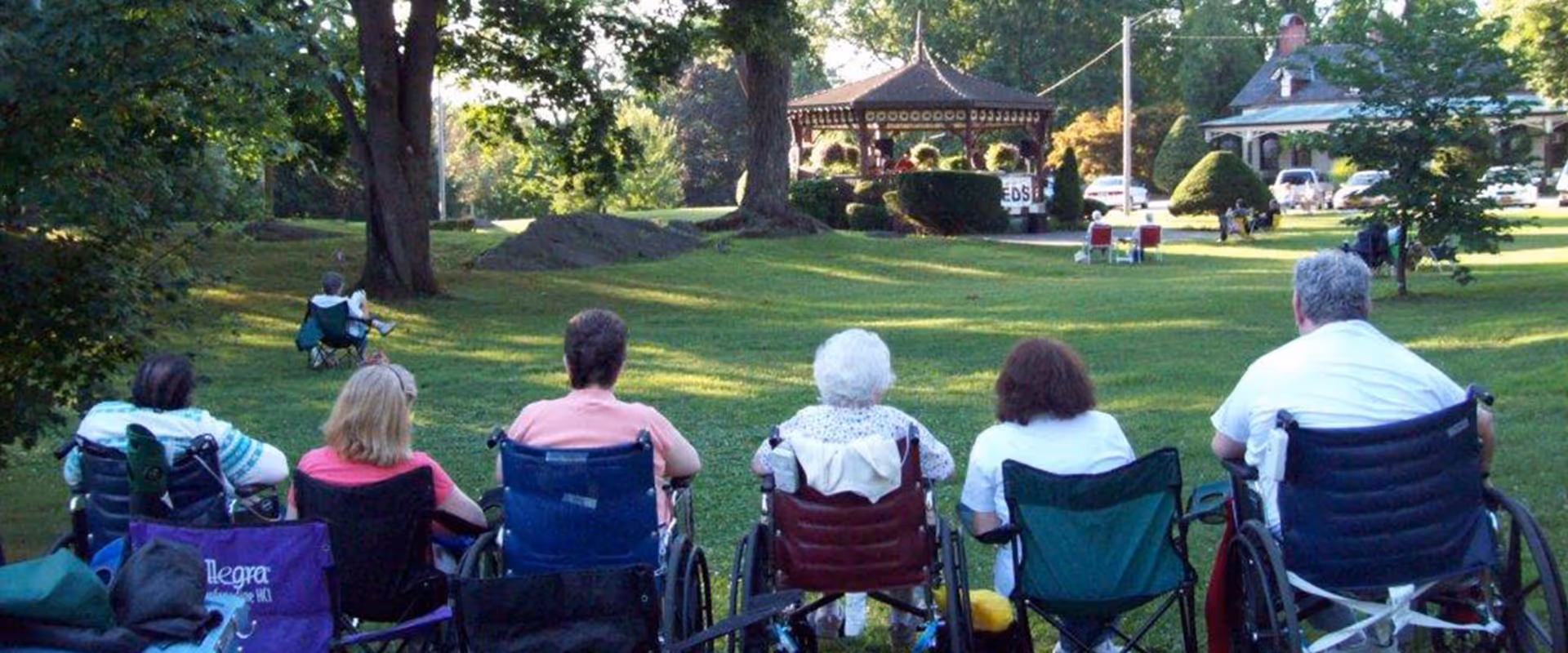 A row of seniors in wheelchairs and lawn chairs sit on a grassy lawn facing a gazebo in a park-like setting.