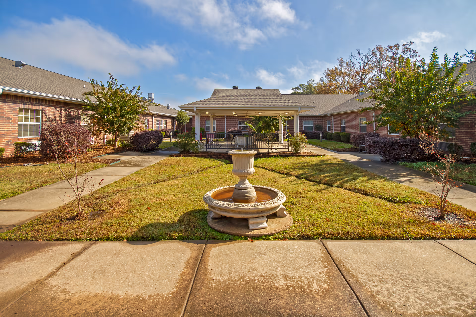 Outdoor courtyard area of a senior living facility with a central stone fountain surrounded by grass and paved walkways. The courtyard is bordered by single-story brick buildings with windows and a covered patio area with seating. Trees and shrubs are planted around the courtyard under a partly cloudy blue sky.