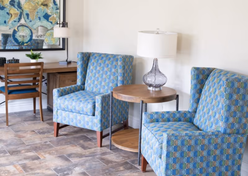 Two patterned blue armchairs flank a round wooden side table with a lamp in a bright seating area with a desk and map on the wall.