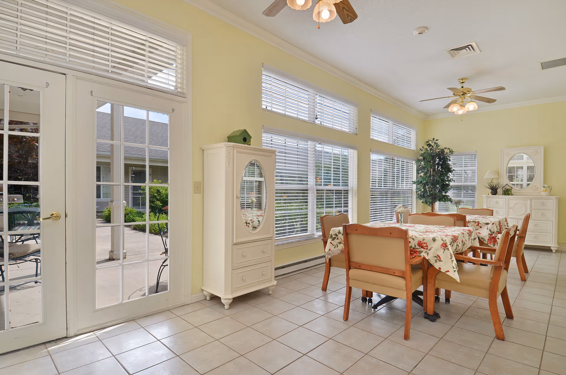 A bright dining room with large windows and glass doors leading outside. The room features a table covered with a floral tablecloth surrounded by six wooden chairs with cushioned seats. There are two ceiling fans with lights, a white cabinet with an oval mirror, a white dresser with a round mirror, and a tall potted plant in the corner.