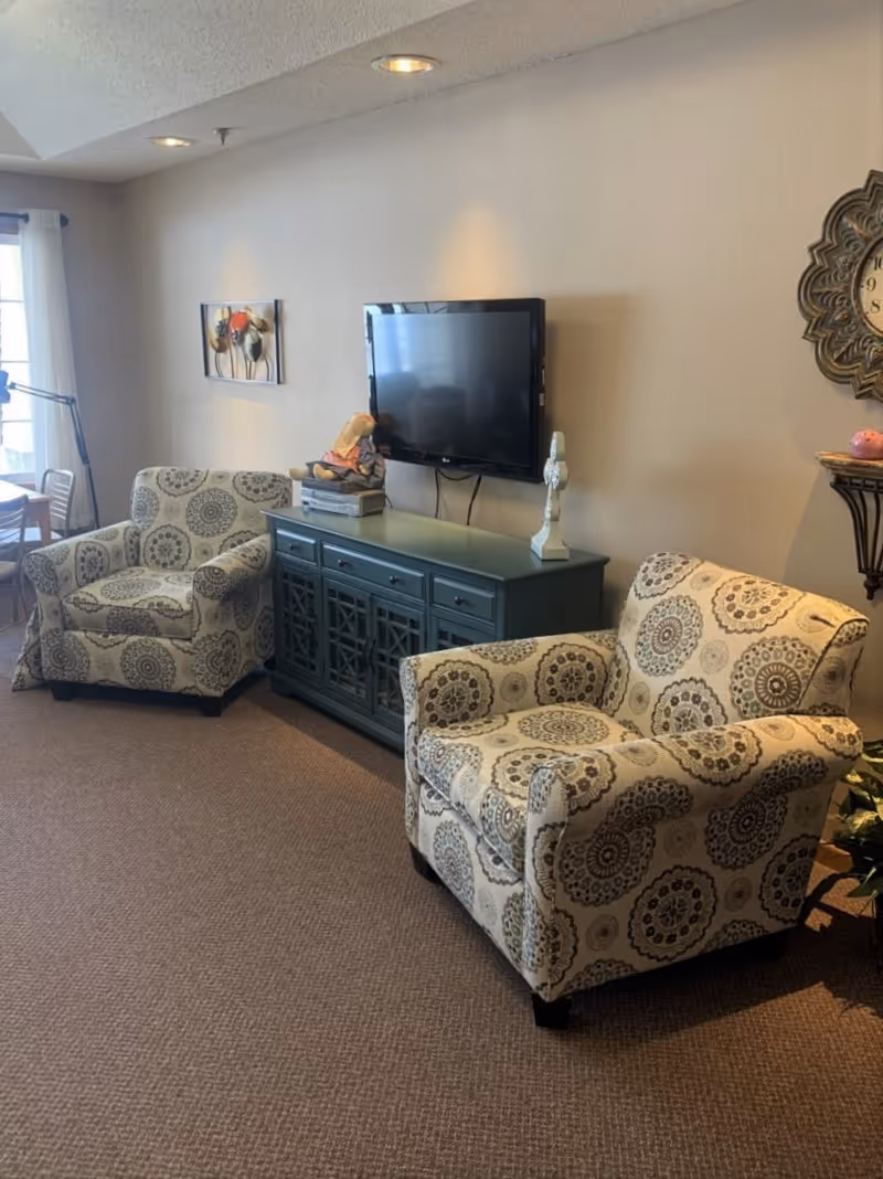 A cozy living room area with two patterned armchairs facing a wall-mounted flat screen TV above a blue-green cabinet. The room has beige walls, a carpeted floor, a decorative wall clock, and a small table with a lamp and decor items. Natural light comes in from a window with white curtains on the left side.