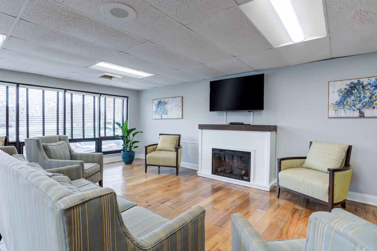 Bright senior living common room with upholstered chairs arranged around a fireplace and wall-mounted TV and large windows along one wall.