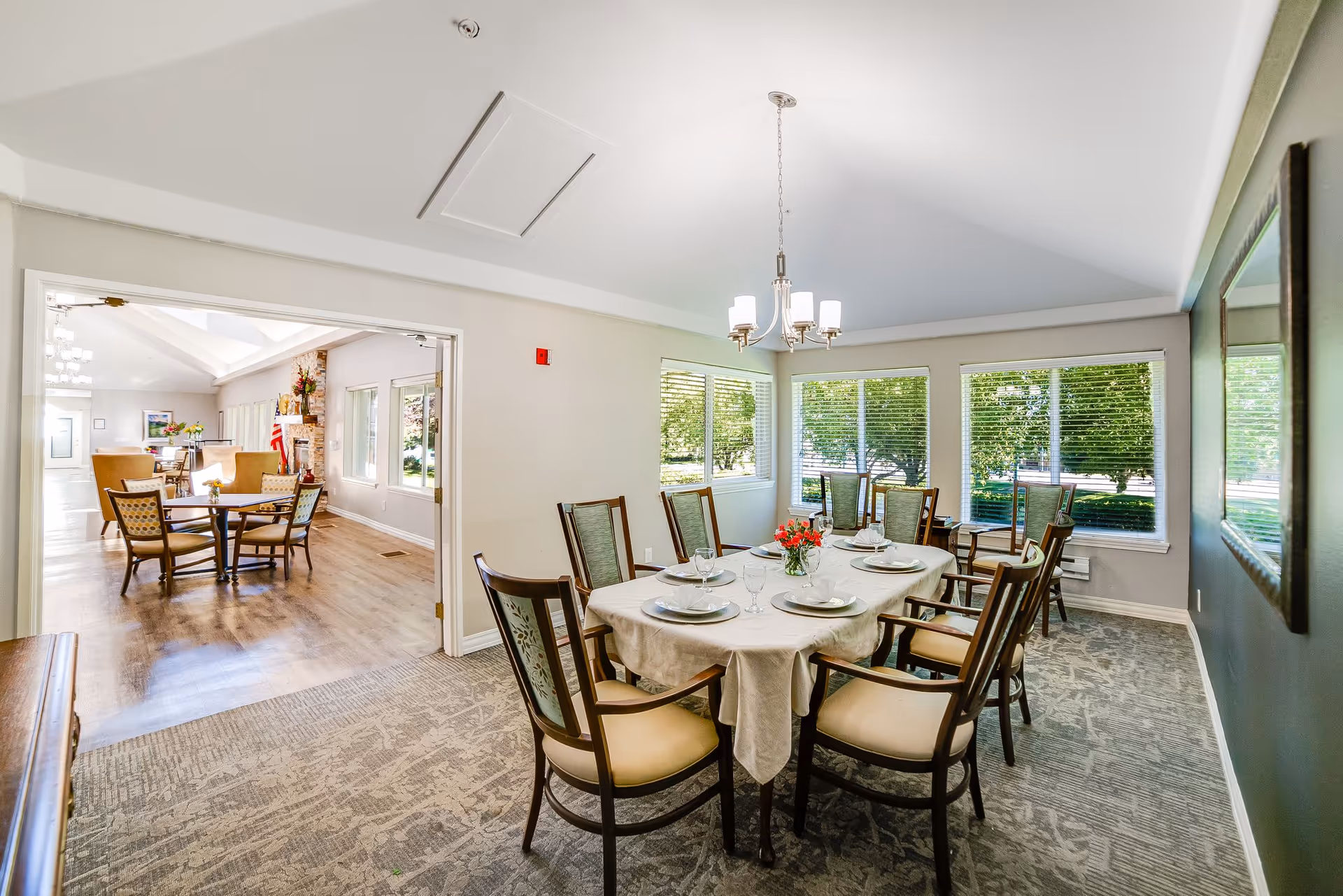 A bright dining room set for a meal with a round table and eight chairs, chandelier, large windows overlooking greenery, and an adjacent sitting area visible through a doorway.