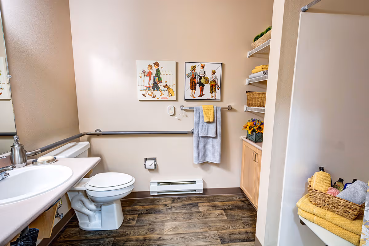 A clean and accessible bathroom with a white toilet, a sink with a soap dispenser, and grab bars along the walls. There are two framed pictures of children on the wall above a towel rack holding a gray and a yellow towel. Shelves with neatly folded towels and a basket with flowers are visible on the right side. The floor has wood-like vinyl flooring.