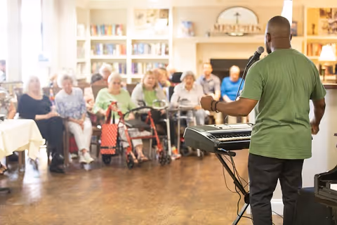 A man in a green shirt stands and sings into a microphone while playing a keyboard in front of a seated audience of elderly people in a well-lit room with bookshelves and a fireplace.