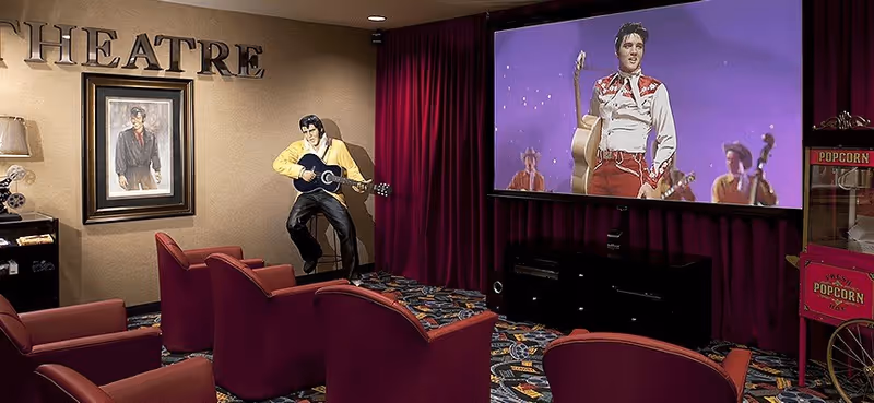 Small theater-style room with red upholstered seats facing a large screen showing a performer, a life-size performer statue with a guitar, framed artwork, and a popcorn machine.
