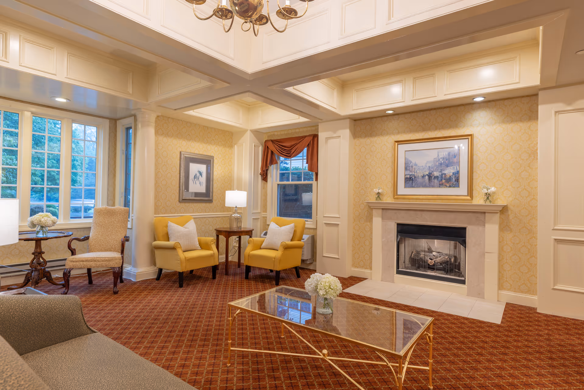 A cozy living room with a fireplace, two yellow armchairs with white pillows, a glass coffee table with a flower vase, a beige sofa, and large windows letting in natural light. The walls are decorated with framed artwork and patterned wallpaper, and the room has a warm, inviting atmosphere.