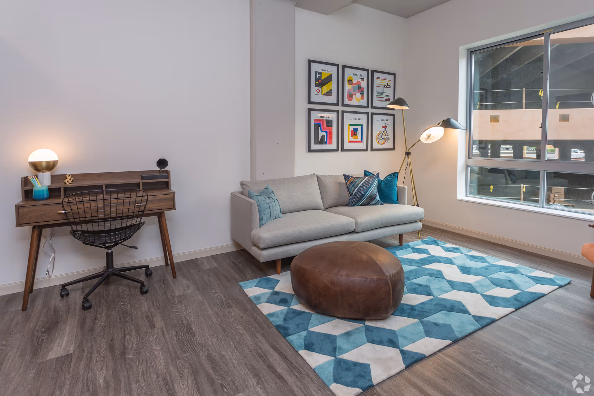 A modern living room with a light gray sofa adorned with blue and patterned pillows, a brown leather ottoman on a blue and white geometric rug, a wooden desk with a black wire chair and a round lamp, a floor lamp with two lights, and six framed abstract art pieces on the wall. A large window shows an exterior view of a building under construction.