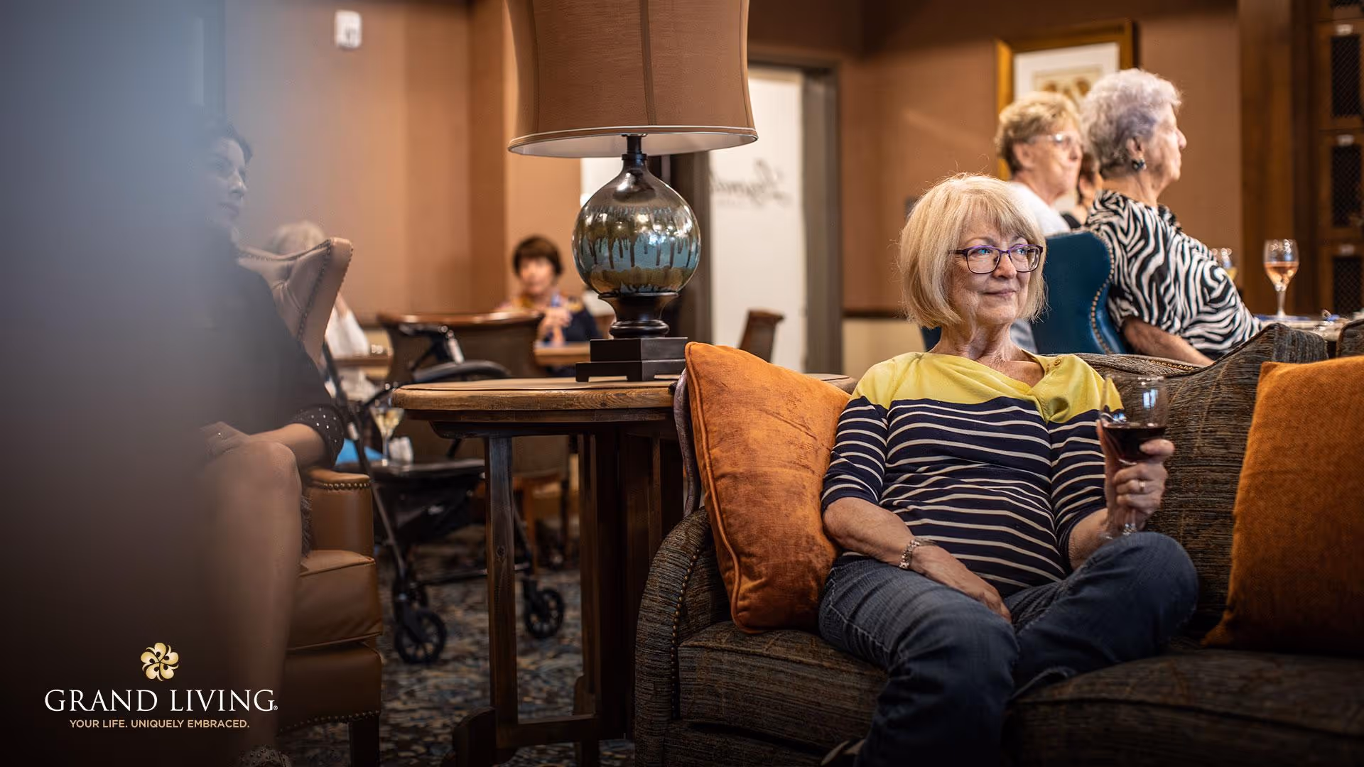 A group of elderly women sitting and relaxing in a warmly decorated living room area. One woman in the foreground is seated on a couch holding a glass of red wine, while others are seated in chairs in the background. A table with a lamp is positioned between the seating areas.