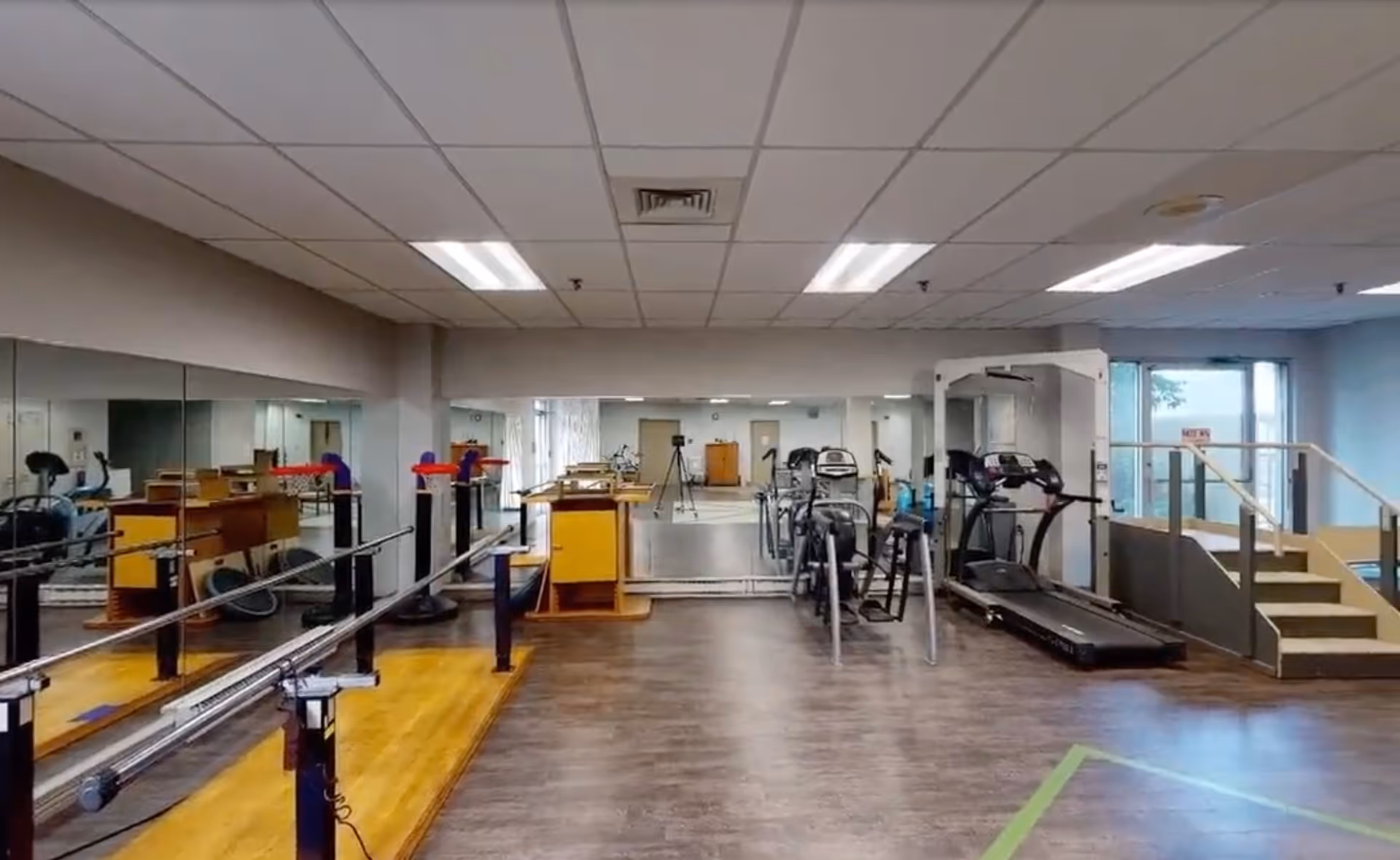 A spacious exercise room with wooden and laminate flooring, featuring parallel bars for walking support, several exercise machines including treadmills and stationary bikes, large mirrors on the walls, and a set of stairs with handrails leading to a raised platform near a glass door.