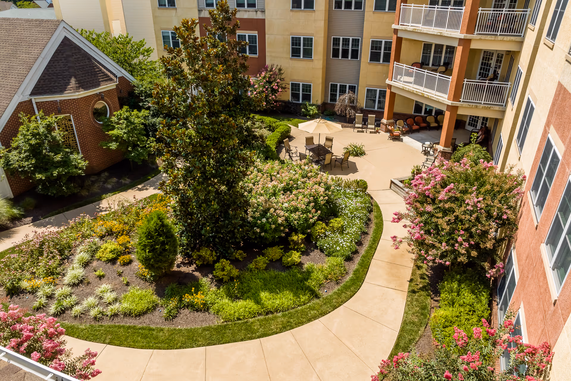 A landscaped outdoor courtyard area at Lutheran Community at Telford featuring a curved walkway, various flowering plants and shrubs, a large tree, and a patio with tables, chairs, and umbrellas. Surrounding buildings with multiple windows and balconies are visible.