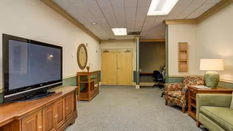 Lounge area in a senior living facility with TV, seating, and a hallway.
