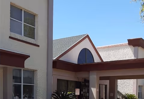 Exterior view of a senior living facility building with beige walls, multiple windows, and a covered entrance with a triangular roof section. There are some plants near the entrance and a clear blue sky above.