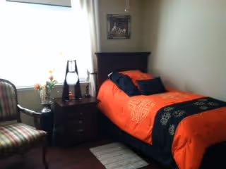 A small bedroom with a single dark wooden bed featuring an orange and black bedspread and a black pillow. Next to the bed is a dark wooden nightstand with a lamp and decorative items. A striped upholstered chair is positioned near a window with white curtains, and a framed picture hangs on the wall above the bed.