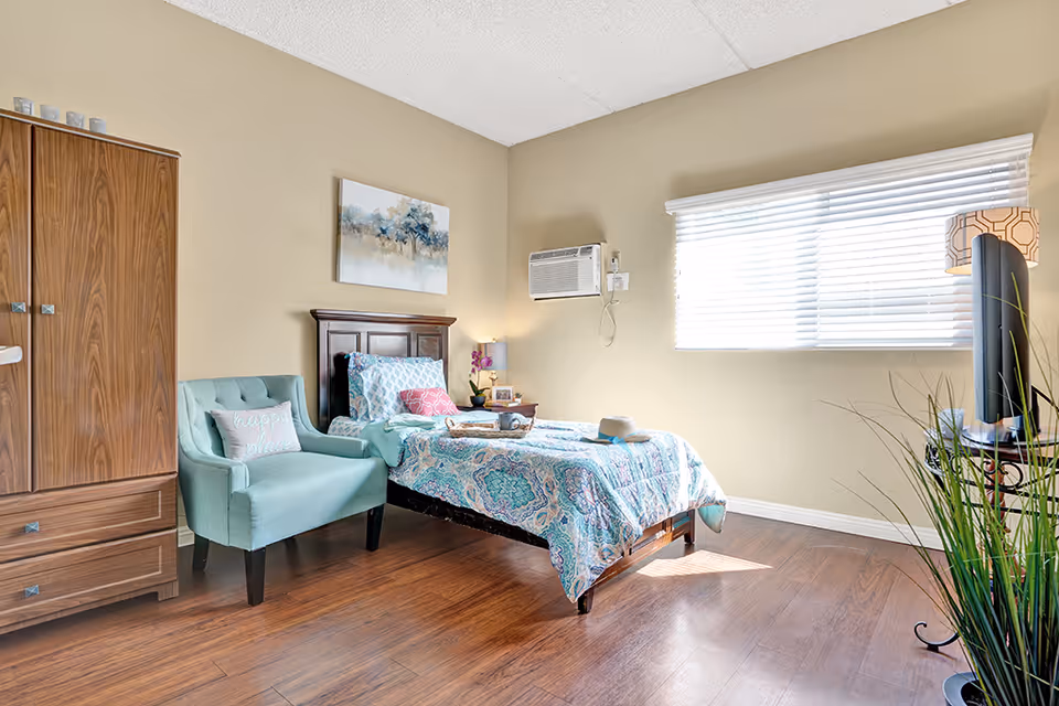 A bright and cozy bedroom with a single bed covered in a blue and white patterned comforter. Next to the bed is a small wooden nightstand with a lamp and a small plant. A light blue armchair with a pillow that says 'happy place' is positioned beside a wooden wardrobe. The room has wooden flooring, beige walls, a window with white blinds, and a wall-mounted air conditioning unit.