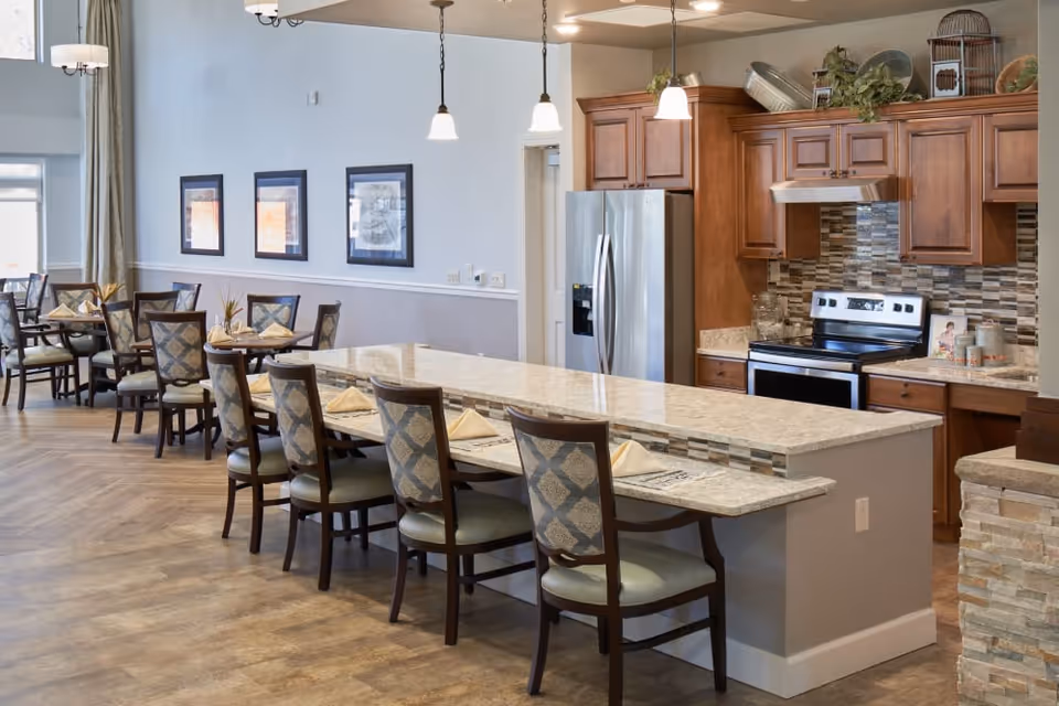 A spacious dining area with a long granite countertop bar and several chairs with patterned upholstery. Behind the bar is a kitchen area with wooden cabinets, a stainless steel refrigerator, stove, and a tiled backsplash. Additional dining tables and chairs are visible in the background along the wall with framed pictures.