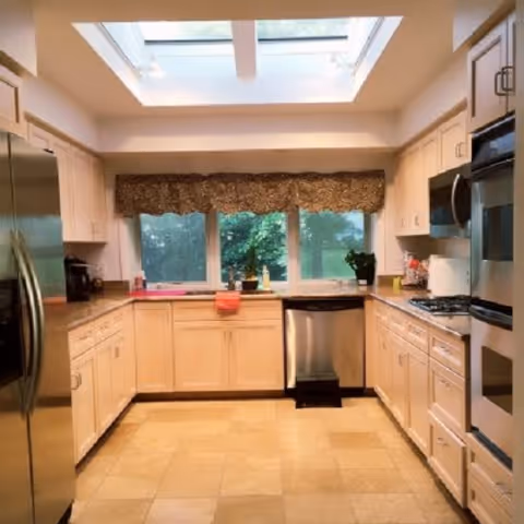 Bright kitchen with beige cabinets, stainless steel refrigerator, dishwasher, oven, and microwave. Large windows with a decorative valance let in natural light from a skylight above. The countertops have small plants and kitchen items.