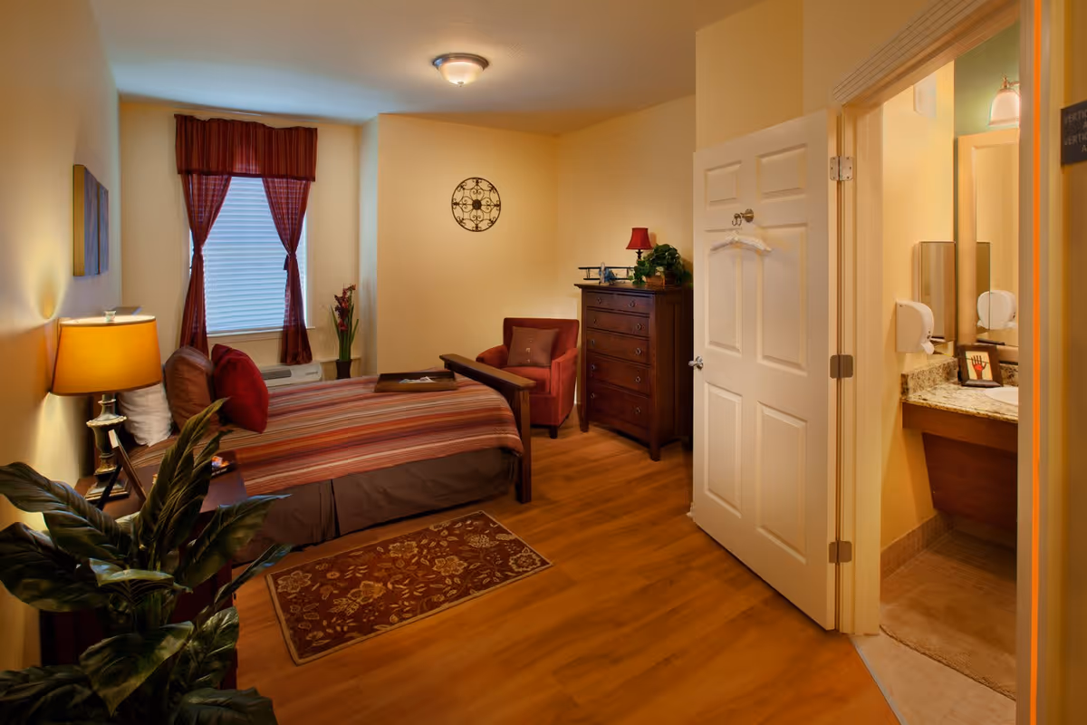 A cozy bedroom with a bed covered in a striped bedspread, a bedside table with a lamp, a red armchair, a wooden dresser, and a window with red curtains. The room has wooden flooring and warm yellow walls. An open door reveals a bathroom with a granite countertop and a mirror.