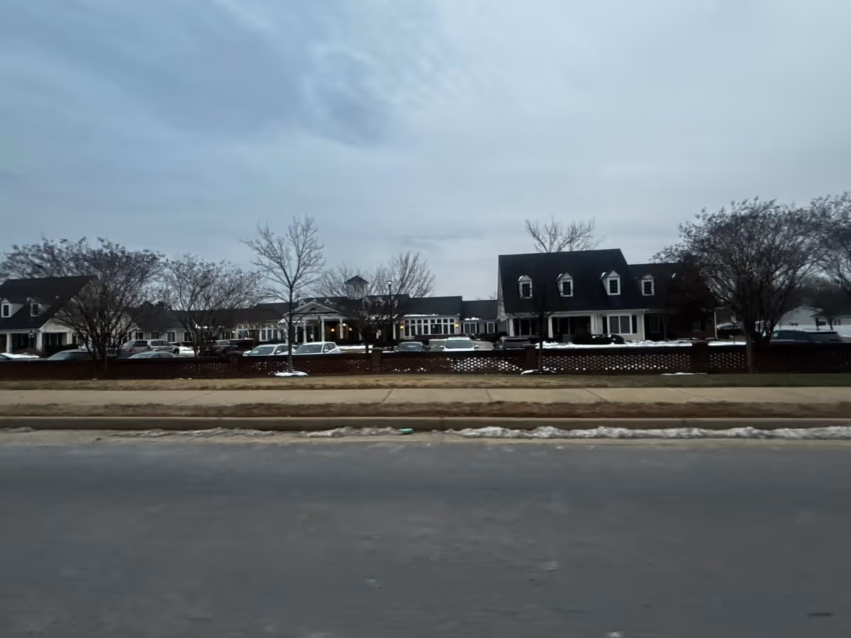Front exterior of a long senior living building with dormer windows, parked cars, leafless trees and a low brick fence along the road.
