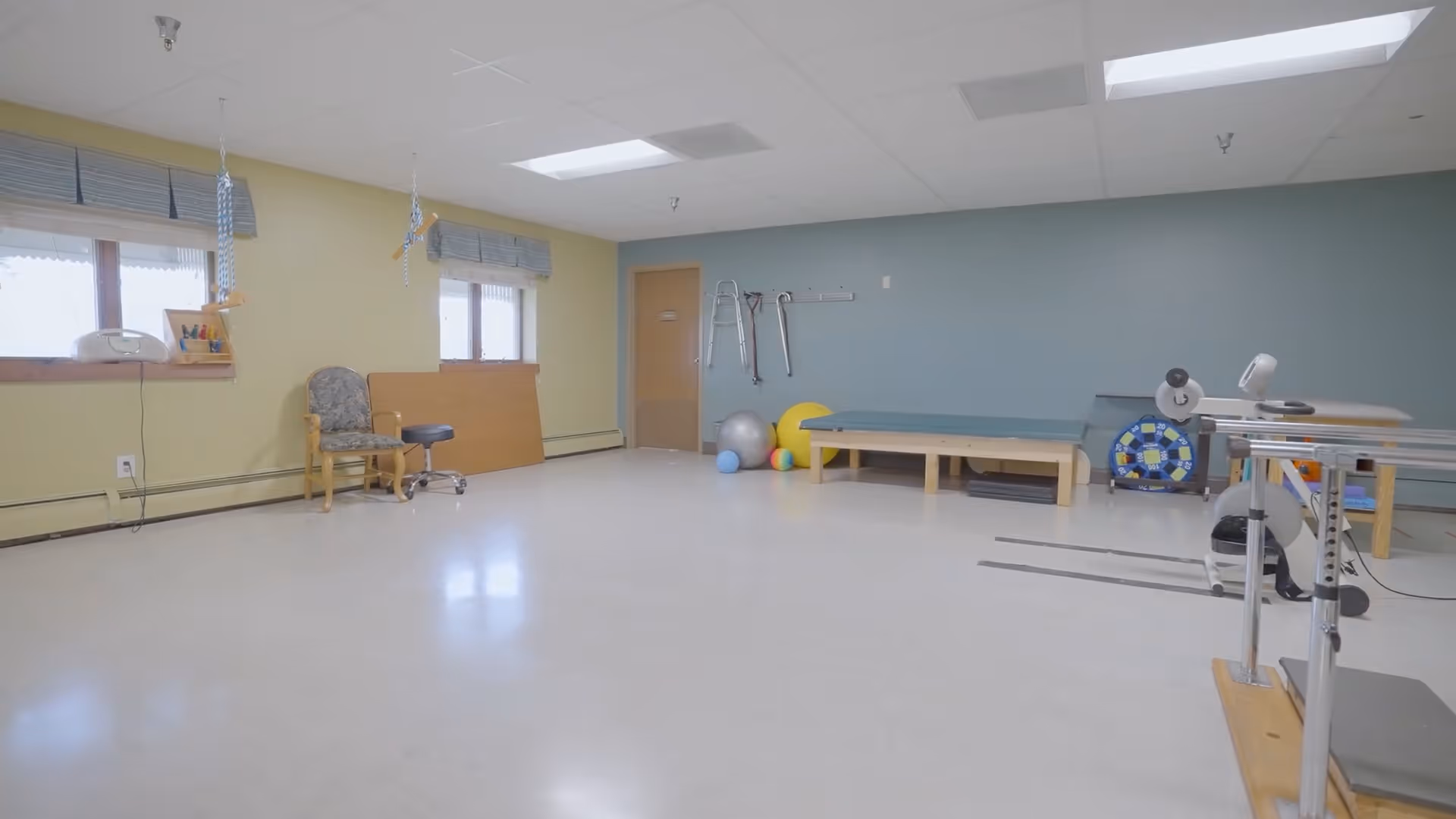 A spacious physical therapy or rehabilitation room with exercise equipment including therapy balls, parallel bars, a treatment table, and various mobility aids hanging on the wall. The room has light yellow and blue walls, two windows with blinds, and a tiled floor.
