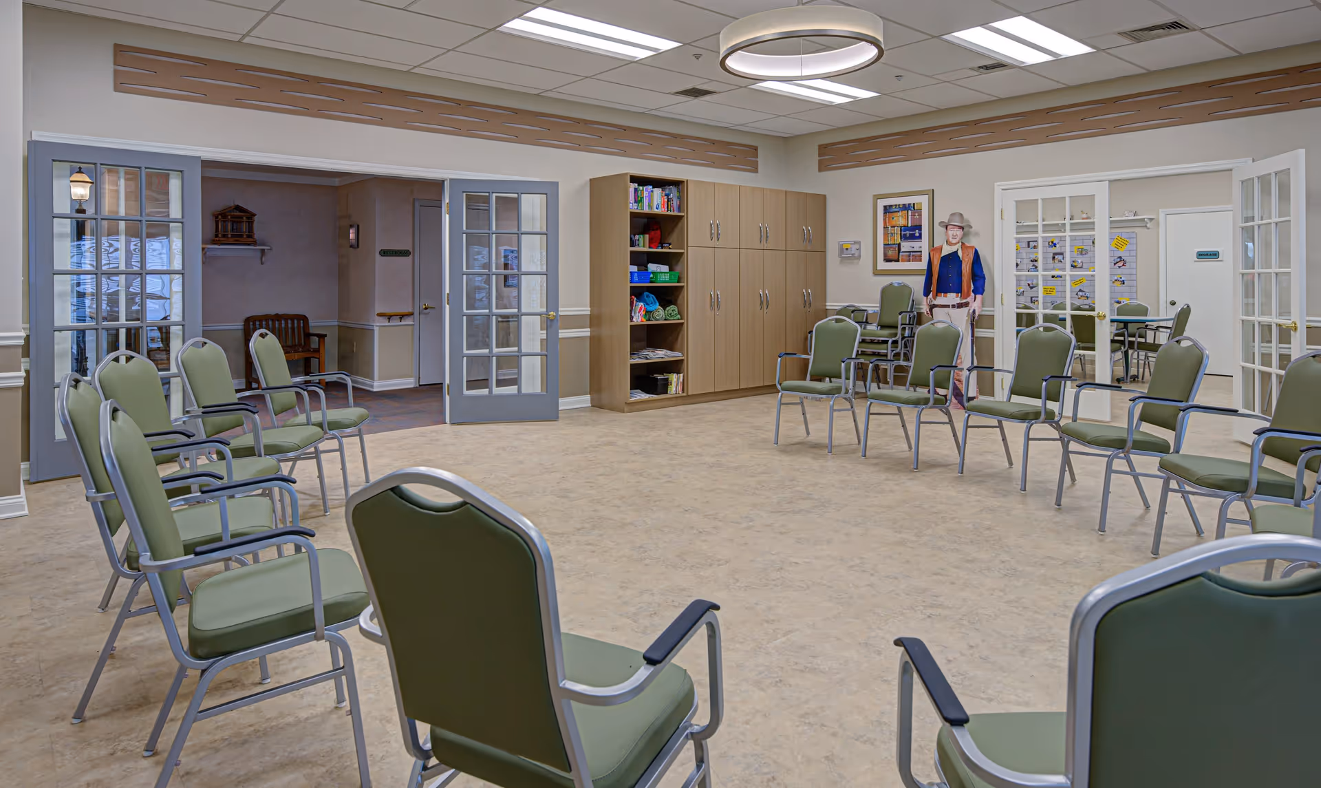 A spacious room with green cushioned chairs arranged in a large circle on a beige floor. The room has light-colored walls with wooden accents near the ceiling and a modern circular light fixture. There are double glass doors on two sides, wooden cabinets with shelves holding various items, and a life-sized cutout figure of a person wearing a hat and vest near the back wall. Another room with tables and chairs is visible through the glass doors.