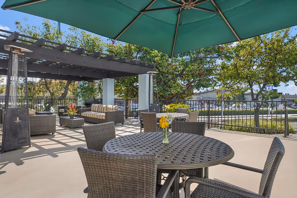 Outdoor senior living patio with a round metal table and wicker chairs under a large green umbrella and a pergola-covered seating area.