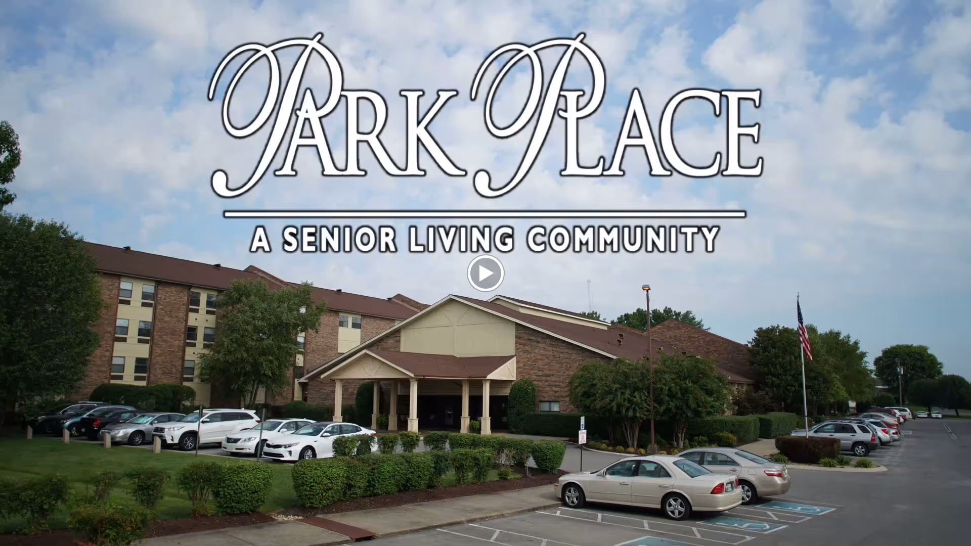 Front exterior of Park Place senior living building with entrance canopy, landscaped grounds and a parking lot with cars.