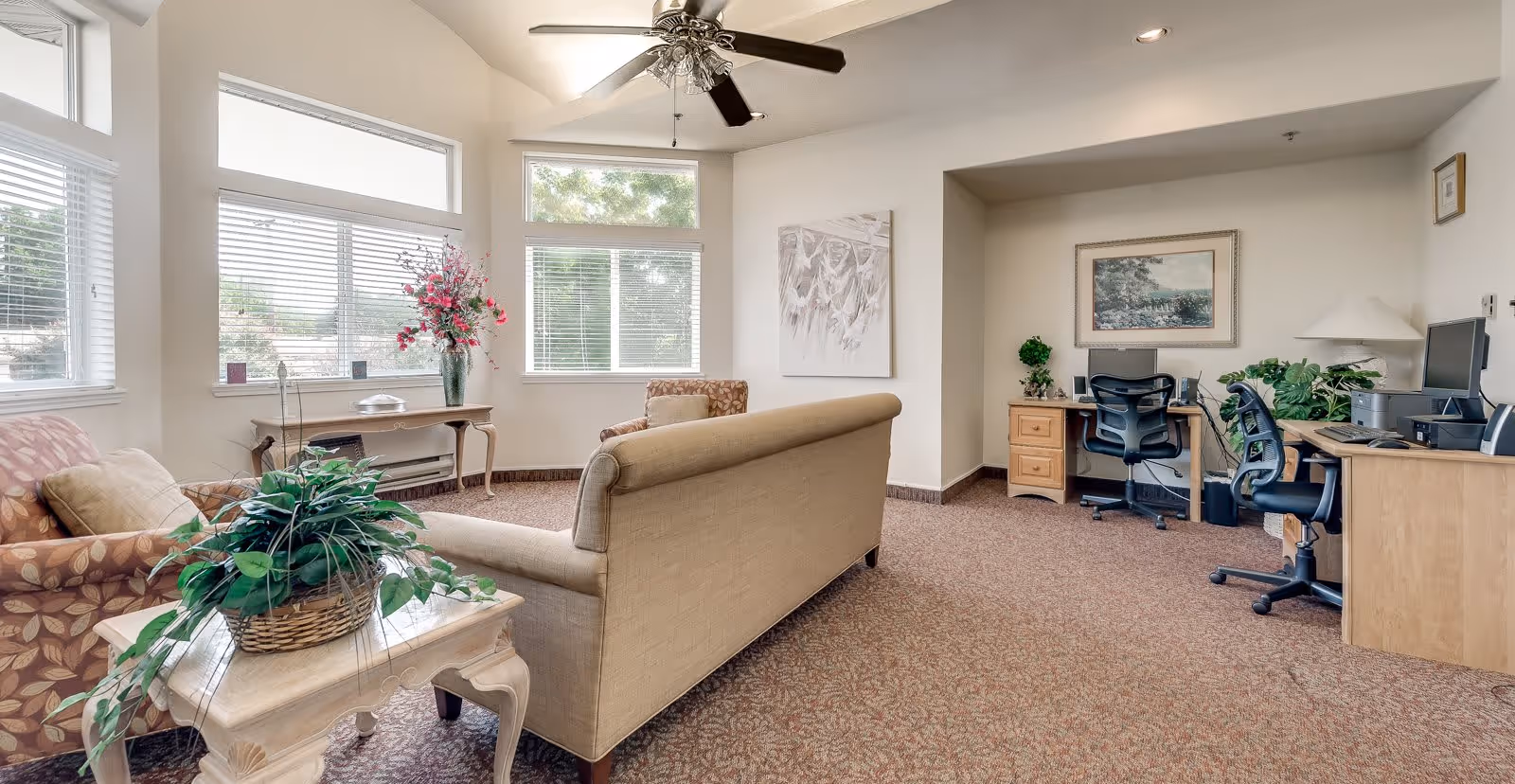 A bright and spacious living room area with large windows letting in natural light. The room features a beige sofa, two patterned armchairs, a small wooden table with a basket of green plants, and a console table with a vase of pink flowers. On the right side, there is a workspace with two desks, office chairs, computers, and a lamp. The walls are decorated with framed artwork and the ceiling has a fan with lights.