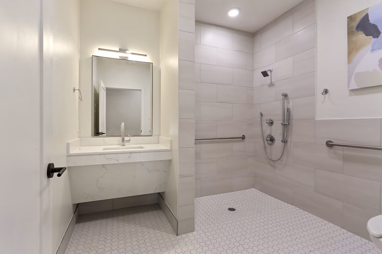 Accessible modern bathroom with a wall-mounted sink and mirror beside a roll-in tiled shower with grab bars and a handheld showerhead.